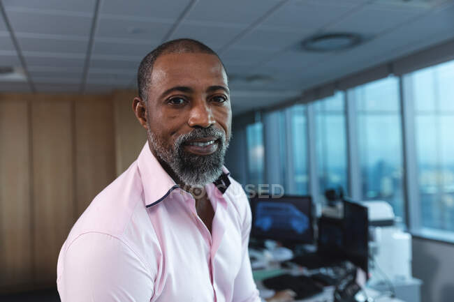 Retrato de un hombre de negocios afroamericano trabajando hasta tarde en una oficina moderna, mirando a la cámara y sonriendo. - foto de stock