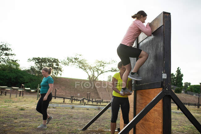 Gruppo multietnico di donne che indossano magliette colorate in una sessione di allenamento di boot camp, esercitandosi, arrampicandosi su una parete e aiutandosi a vicenda. Esercizio di gruppo all'aperto, divertente sfida sana. — Foto stock