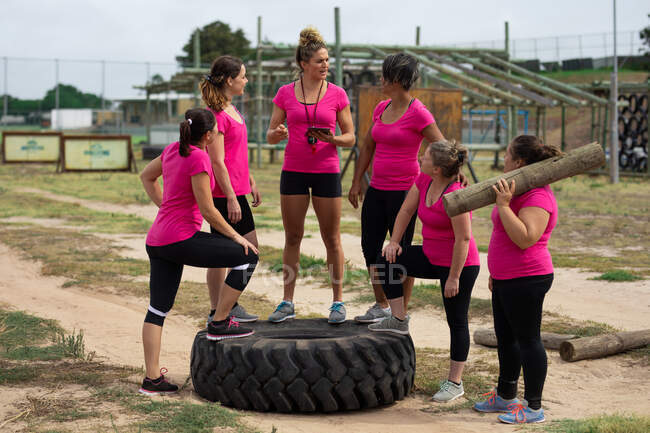 Grupo multiétnico de mujeres todas con camisetas rosas en una sesión de entrenamiento en el campo de entrenamiento, haciendo ejercicio y motivándolas en su sofá. Ejercicio en grupo al aire libre, divertido desafío saludable. - foto de stock