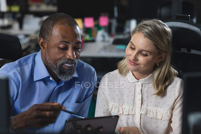 Кавказская деловая женщина и афроамериканский бизнесмен, работающие допоздна в современном офисе, сидящие за столом, за планшетным компьютером, обсуждающие свою работу. — стоковое фото