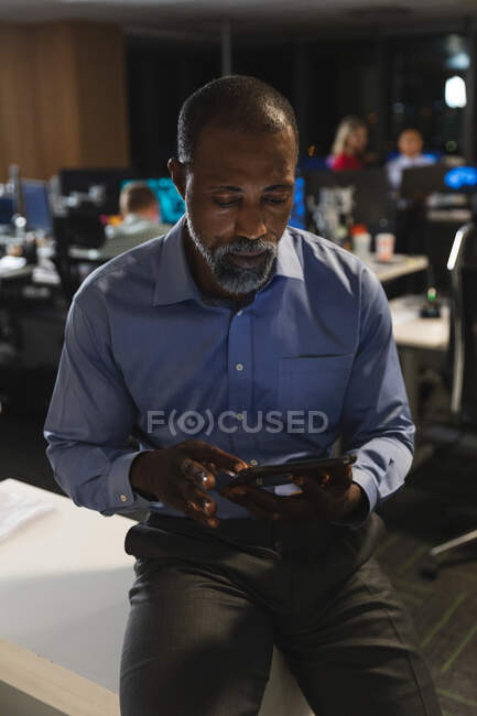 Uomo d'affari afroamericano che lavora fino a tardi la sera in un ufficio moderno, seduto su una scrivania, usando un tablet. — Foto stock