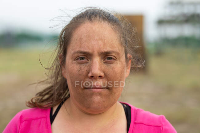 Portrait of a mixed race woman wearing pink t shirt at a boot camp training session, exercising, posing for a photo with dirt on her face. Outdoor group exercise, fun healthy challenge. — Stock Photo