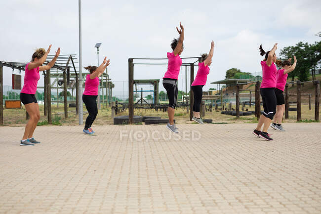 Grupo multi-étnico de mulheres todas vestindo camisetas cor-de-rosa em uma sessão de treinamento de campo de treinamento, exercitando-se, fazendo macacos de salto. Exercício de grupo ao ar livre, desafio saudável divertido. — Fotografia de Stock