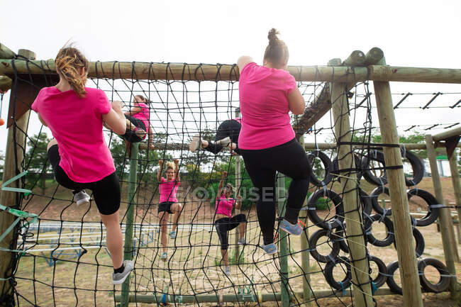 Grupo multi-étnico de mulheres todas vestindo camisetas cor-de-rosa em uma sessão de treinamento de campo de treinamento, exercitando-se, escalando em redes sobre uma estrutura de escalada. Exercício de grupo ao ar livre, desafio saudável divertido. — Fotografia de Stock