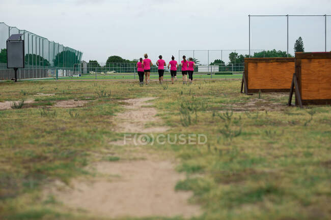 Groupe multiethnique de femmes portant toutes des t-shirts roses lors d'une séance d'entraînement au camp d'entraînement, faisant de l'exercice, courant sur un terrain. Exercice de groupe en plein air, défi sain amusant. — Photo de stock