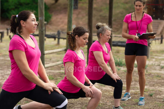 Multi-ethnic group of women all wearing pink t shirts at a boot camp training session, exercising, stretching their legs and couch motivating them. Outdoor group exercise, fun healthy challenge. — Stock Photo