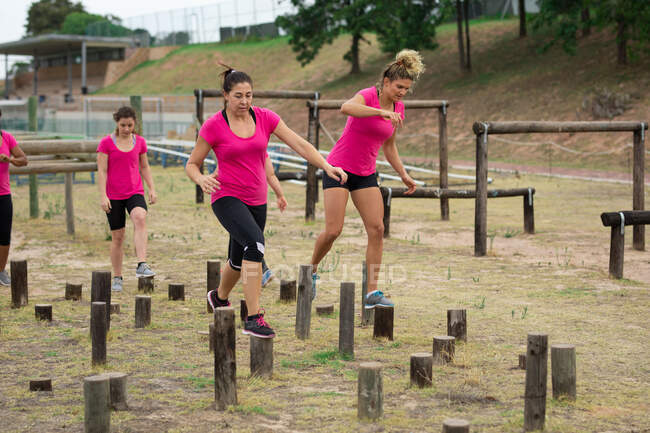 Grupo multi-étnico de mulheres todas vestindo camisetas rosa em uma sessão de treinamento de campo de treinamento, exercitando, equilibrando e andando através de logs. Exercício de grupo ao ar livre, desafio saudável divertido. — Fotografia de Stock