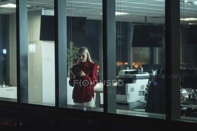 Caucasian businesswoman working late in the evening in a modern office, standing next to a window, using a tablet computer. — Stock Photo