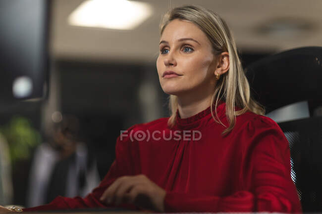 Mujer de negocios caucásica que trabaja hasta tarde en la noche en una oficina moderna, sentado en un escritorio, utilizando una computadora de escritorio. - foto de stock