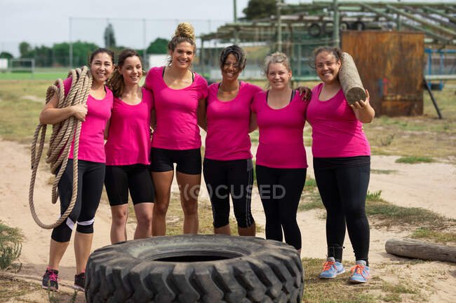Retrato de mujeres mestizas confiadas en un campo de entrenamiento con camisetas rosas posando para una foto con un neumático frente a ellas. Ejercicio en grupo al aire libre, divertido desafío saludable. - foto de stock
