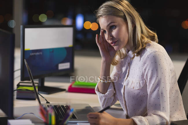 Mujer de negocios caucásica trabajando hasta tarde en la noche en una oficina moderna, sentada en un escritorio, usando una computadora portátil, tomando notas. - foto de stock