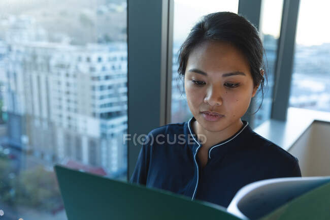 Asiatische Geschäftsfrau arbeitet spät abends in einem modernen Büro, steht am Fenster und blickt auf einen Ordner. — Stockfoto
