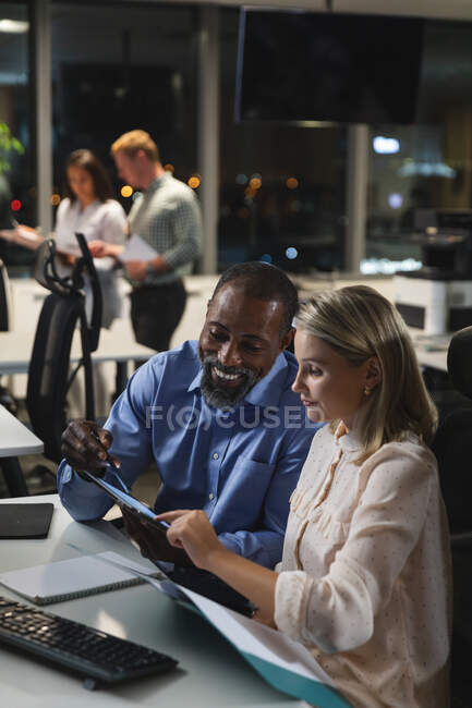 Кавказская деловая женщина и афроамериканский бизнесмен, работающие допоздна в современном офисе, сидящие за столом, за планшетным компьютером, обсуждающие свою работу. — стоковое фото
