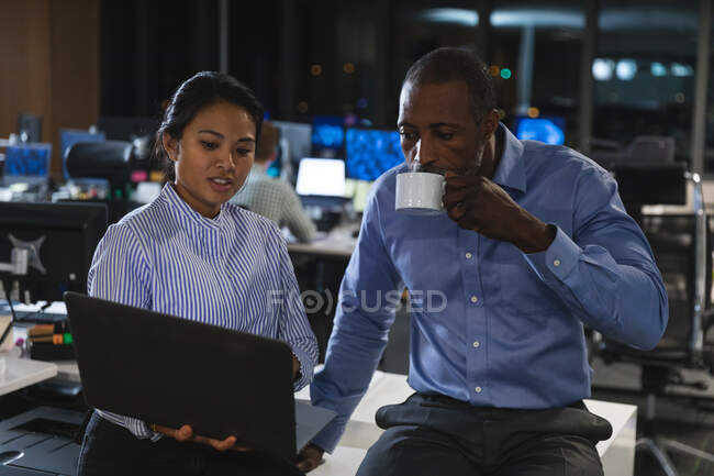 Азиатская деловая женщина и афроамериканский бизнесмен, работающий допоздна в современном офисе, сидящий на столе, пользующийся ноутбуком, мужчина, держащий чашку и пьющий. — стоковое фото