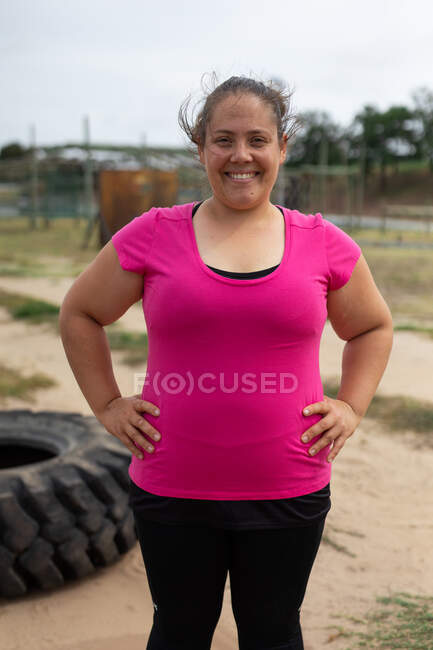 Retrato de una mujer de raza mixta segura y feliz en un campamento de entrenamiento, usando una camiseta rosa, un neumático en el fondo. Ejercicio en grupo al aire libre, divertido desafío saludable. - foto de stock