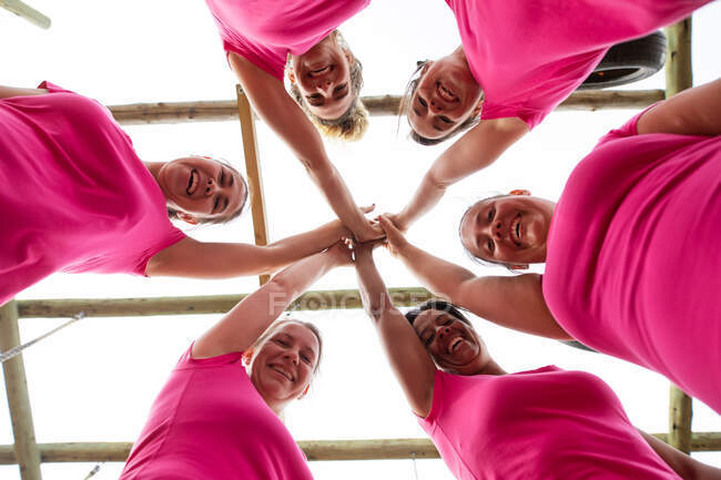 Grupo multiétnico de mujeres que usan camisetas rosas en una sesión de entrenamiento de campo de entrenamiento, ejercitándose, motivándose mutuamente y apilándose las manos. Ejercicio en grupo al aire libre, divertido desafío saludable. - foto de stock