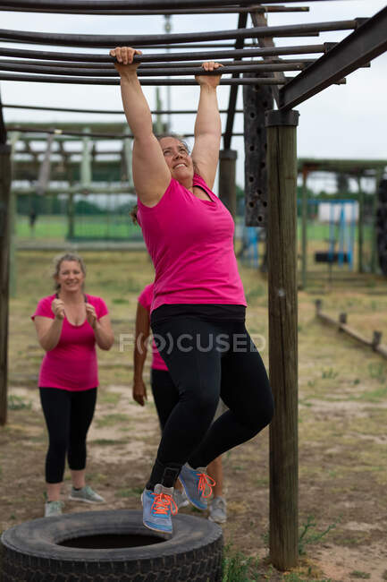 Grupo multi-étnico de mulheres todas vestindo camisetas cor-de-rosa em uma sessão de treinamento de campo de treinamento, exercício, pendurado em bares de macacos. Exercício de grupo ao ar livre, desafio saudável divertido. — Fotografia de Stock