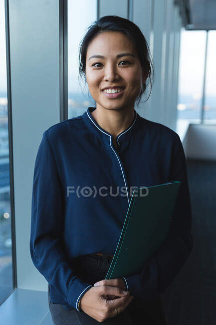 Porträt einer asiatischen Geschäftsfrau, die spät am Abend in einem modernen Büro arbeitet, neben einem Fenster steht, einen Ordner in der Hand hält, in die Kamera blickt und lächelt. — Stockfoto
