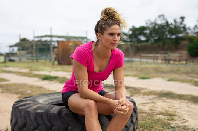 Una donna caucasica sicura di sé in un campo di addestramento per una sessione di allenamento, indossando una maglietta rosa, seduta su una grande gomma. Esercizio di gruppo all'aperto, divertente sfida sana. — Foto stock