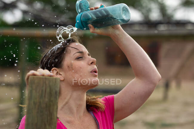 Une femme caucasienne portant un t-shirt rose lors d'une séance d'entraînement au camp d'entraînement, faisant de l'exercice, faisant une pause, versant de l'eau sur son visage. Exercice de groupe en plein air, défi sain amusant. — Photo de stock