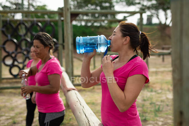 Grupo multi-étnico de mulheres todas vestindo camisetas cor-de-rosa em uma sessão de treinamento de campo de treinamento, exercitando, fazendo uma pausa, bebendo água. Exercício de grupo ao ar livre, desafio saudável divertido. — Fotografia de Stock