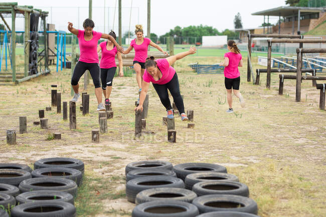 Gruppo multietnico di donne che indossano t-shirt rosa in una sessione di allenamento di boot camp, esercitandosi, bilanciando e camminando tra i tronchi. Esercizio di gruppo all'aperto, divertente sfida sana. — Foto stock