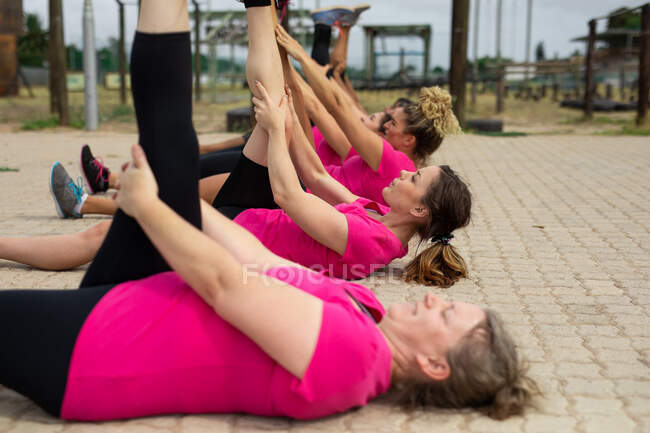 Multi-ethnic group of women all wearing pink t shirts at a boot camp training session, exercising, stretching their legs, lying on the ground. Outdoor group exercise, fun healthy challenge. — Stock Photo
