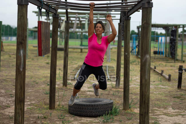 Mujer de raza mixta con camiseta rosa en una sesión de entrenamiento de campo de entrenamiento de arranque, haciendo ejercicio, colgando de barras de mono. Ejercicio en grupo al aire libre, divertido desafío saludable. - foto de stock