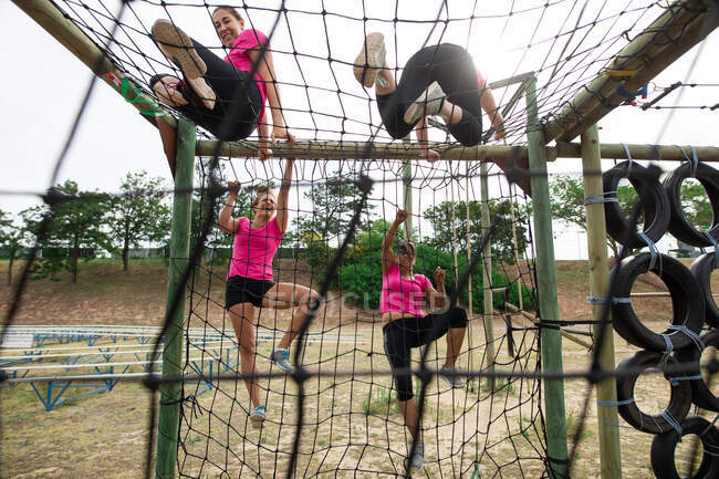 Grupo multiétnico de mujeres todas con camisetas rosas en una sesión de entrenamiento de campo de entrenamiento, ejercicio, escalada en redes sobre un marco de escalada. Ejercicio en grupo al aire libre, divertido desafío saludable. - foto de stock