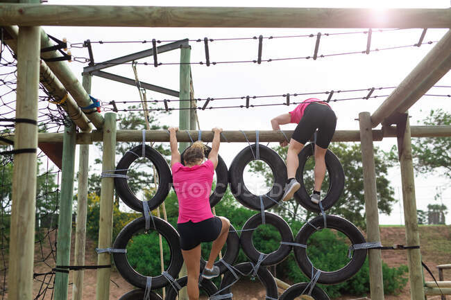 Groupe multiethnique de femmes portant toutes des t-shirts roses lors d'une séance d'entraînement au camp d'entraînement, faisant de l'exercice, escaladant un mur de pneus sur un cadre d'escalade. Exercice de groupe en plein air, défi sain amusant. — Photo de stock