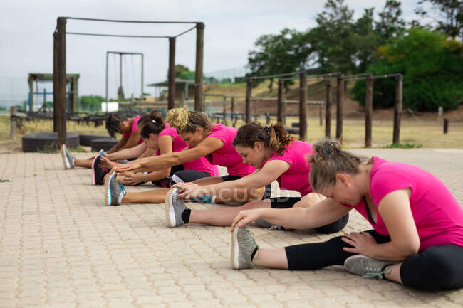 Grupo multiétnico de mujeres todas con camisetas rosas en una sesión de entrenamiento de campo de entrenamiento, haciendo ejercicio, estirando sus piernas, sentadas en el suelo. Ejercicio en grupo al aire libre, divertido desafío saludable. - foto de stock