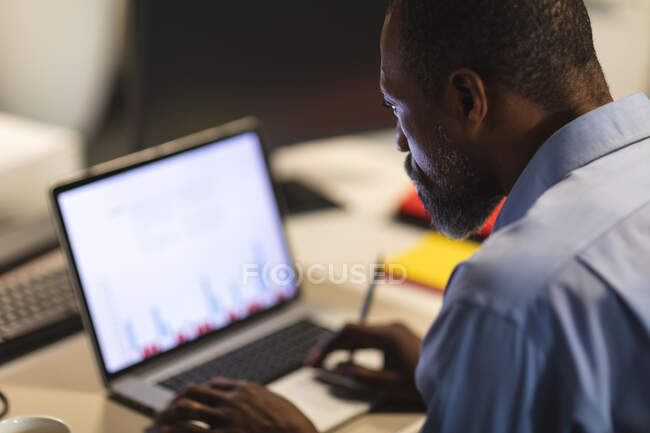 Homme d'affaires afro-américain travaillant tard le soir dans un bureau moderne, assis à un bureau, à l'aide d'un ordinateur portable. — Photo de stock