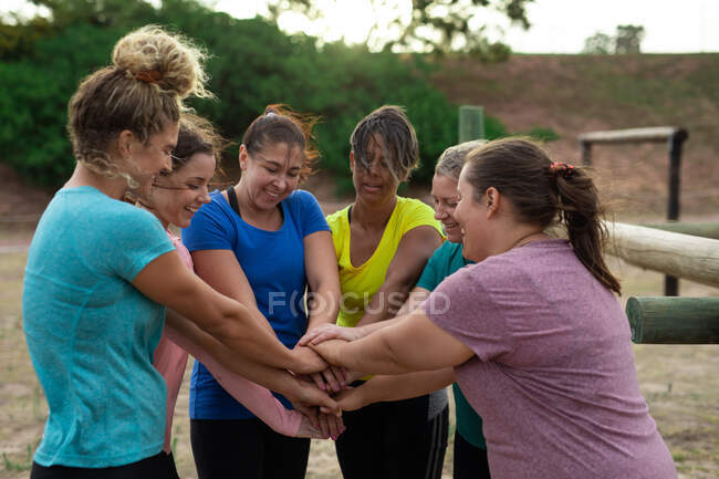 Grupo multi-étnico de mulheres todas vestindo camisetas coloridas em uma sessão de treinamento de campo de treinamento, exercitando, motivando e empilhando mãos. Exercício de grupo ao ar livre, desafio saudável divertido. — Fotografia de Stock