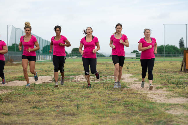 Grupo multiétnico de mujeres todas con camisetas rosas en una sesión de entrenamiento de campo de entrenamiento, ejercicio, correr en un campo. Ejercicio en grupo al aire libre, divertido desafío saludable. - foto de stock