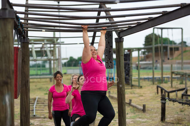 Groupe multiethnique de femmes portant toutes des t-shirts roses lors d'une séance d'entraînement au camp d'entraînement, faisant de l'exercice, suspendues à des barres de singe. Exercice de groupe en plein air, défi sain amusant. — Photo de stock