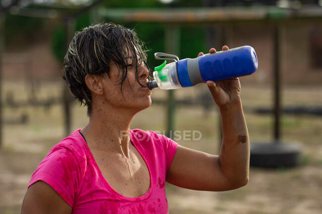 Una mujer de raza mixta vistiendo una camiseta rosa en una sesión de entrenamiento en el campamento de entrenamiento, haciendo ejercicio, tomando un descanso, bebiendo agua. Ejercicio en grupo al aire libre, divertido desafío saludable. - foto de stock