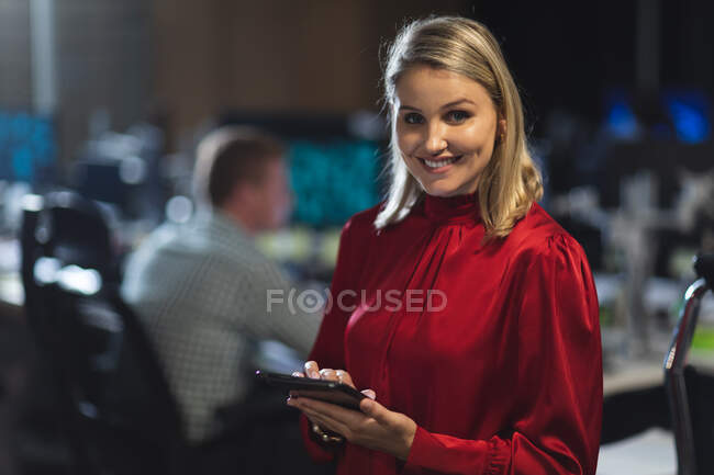Retrato de una mujer de negocios caucásica que trabaja hasta tarde en la noche en una oficina moderna, usando una tableta, mirando a la cámara y sonriendo. - foto de stock