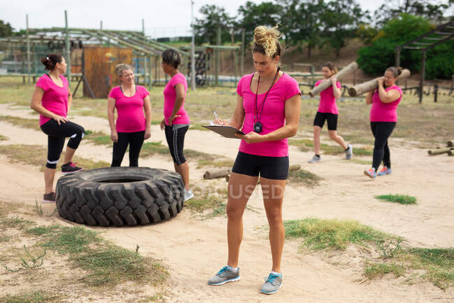 Grupo multi-étnico de mulheres todas vestindo camisetas cor-de-rosa em uma sessão de treinamento de campo de treinamento, exercício e seu sofá segurando um tablet. Exercício de grupo ao ar livre, desafio saudável divertido. — Fotografia de Stock