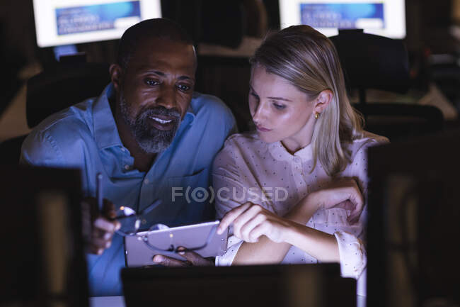 Kaukasische Geschäftsfrau und afroamerikanischer Geschäftsmann arbeiten spät am Abend in einem modernen Büro, sitzen am Schreibtisch, benutzen ein Smartphone und diskutieren über ihre Arbeit. — Stockfoto