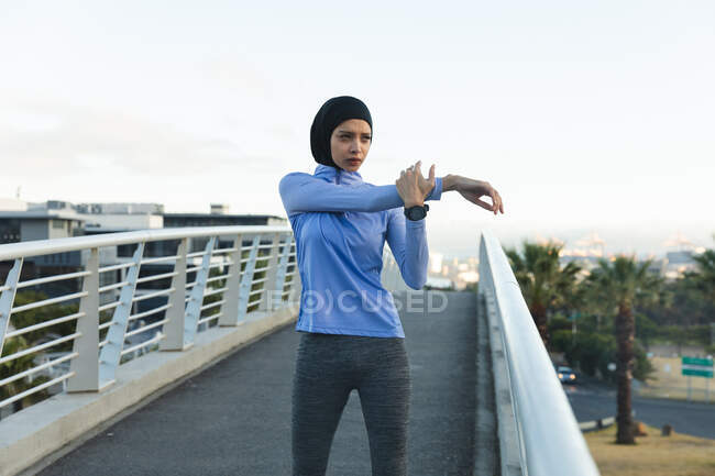 Fit gemischte Rennläuferin in Hijab und Sportbekleidung, die an einem sonnigen Tag im Freien in der Stadt trainiert und ihre Arme auf einer Fußgängerbrücke ausstreckt. Bewegung im städtischen Lebensstil. — Stockfoto
