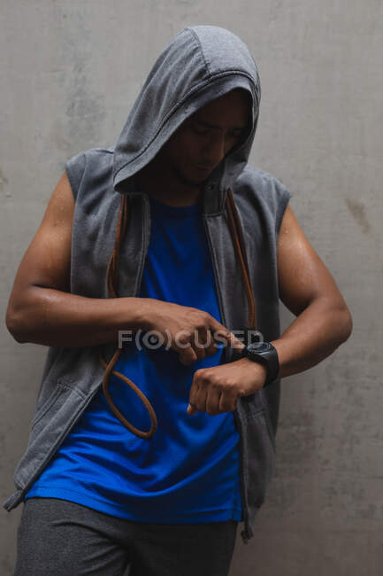 Mixed race man working out in an urban park, wearing hooded top, skipping rope around his neck taking a break checking smartwatch. Fitness strength healthy lifestyle. — Stock Photo