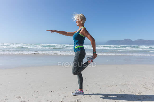 Femme caucasienne senior appréciant l'exercice sur une plage par une journée ensoleillée, pratiquer le yoga, debout en position de yoga avec la mer en arrière-plan. — Photo de stock