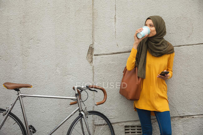 Mischlingshündin in Hijab und gelbem Pullover unterwegs in der Stadt, an der Wand Kaffee zum Mitnehmen trinkend, Smartphone-Fahrrad neben sich haltend. Pendler moderner Lebensstil. — Stockfoto