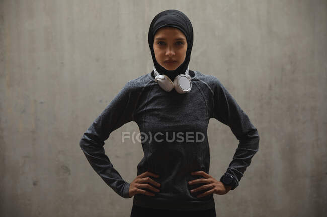 Retrato de mujer de raza mixta en forma usando hijab, auriculares y ropa deportiva ejercitándose al aire libre en la ciudad, mirando a la cámara. Ejercicio urbano. - foto de stock