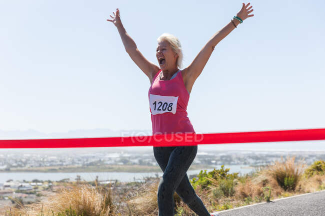 Старша кавказька жінка, яка насолоджується фізичними вправами в сонячний день, святкуючи після бігу і носіння номера, біжить до фінішної лінії.. — стокове фото