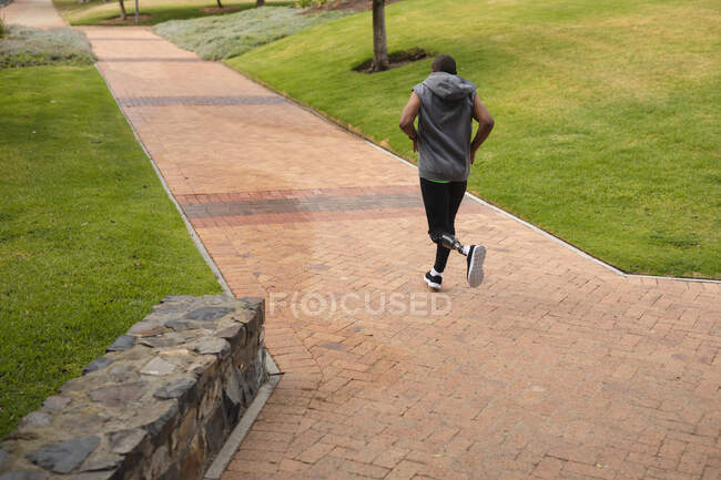 Hombre de raza mixta discapacitado con una pierna protésica, haciendo ejercicio en un parque urbano, con capucha superior corriendo por un camino. Fitness discapacidad estilo de vida saludable. - foto de stock
