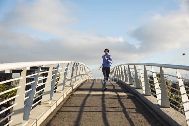 Fit mixed race woman wearing hijab and sportswear exercising outdoors in the city on a sunny day, running on a footbridge. Urban lifestyle exercise. — Stock Photo