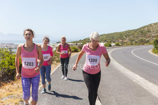 Grupo de amigas caucásicas disfrutando haciendo ejercicio en un día soleado, corriendo, llevando números y ropa deportiva rosa. - foto de stock