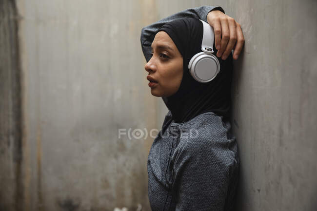 Фіт змішана раса жінка, одягнена в хіджаб, навушники і спортивне лаяння, працює на відкритому повітрі в місті, притулившись до бетонної стіни. Тренування міського життя. — стокове фото