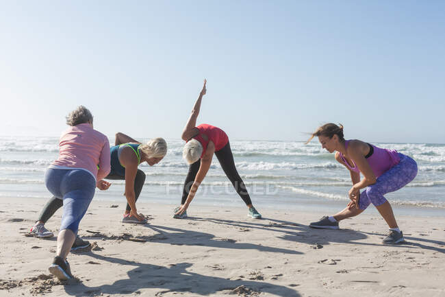 Группа кавказских подруг, занимающихся спортом на пляже в солнечный день, практикующих йогу и растягивающихся на фоне моря. — стоковое фото